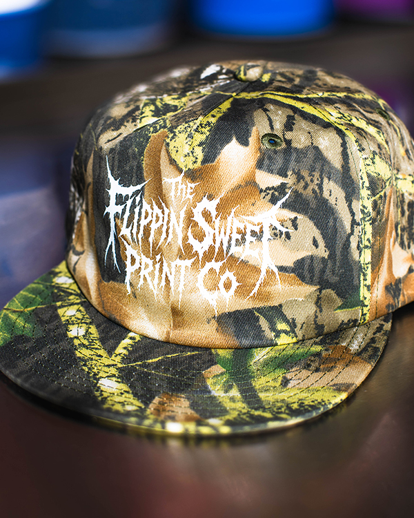 Custom camouflage band cap with white screen printed artwork produced for music merchandise and tour merch.
